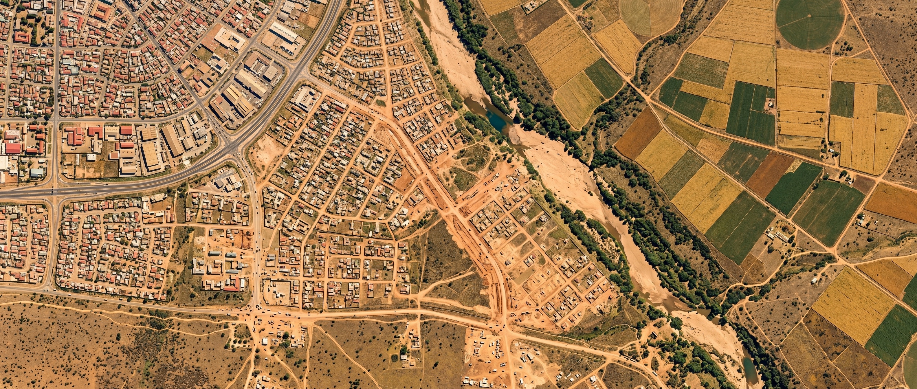 Top-down satellite view of an African city's expanding edge — dense rooftops giving way to dirt-road settlement, a dry riverbed and farmland